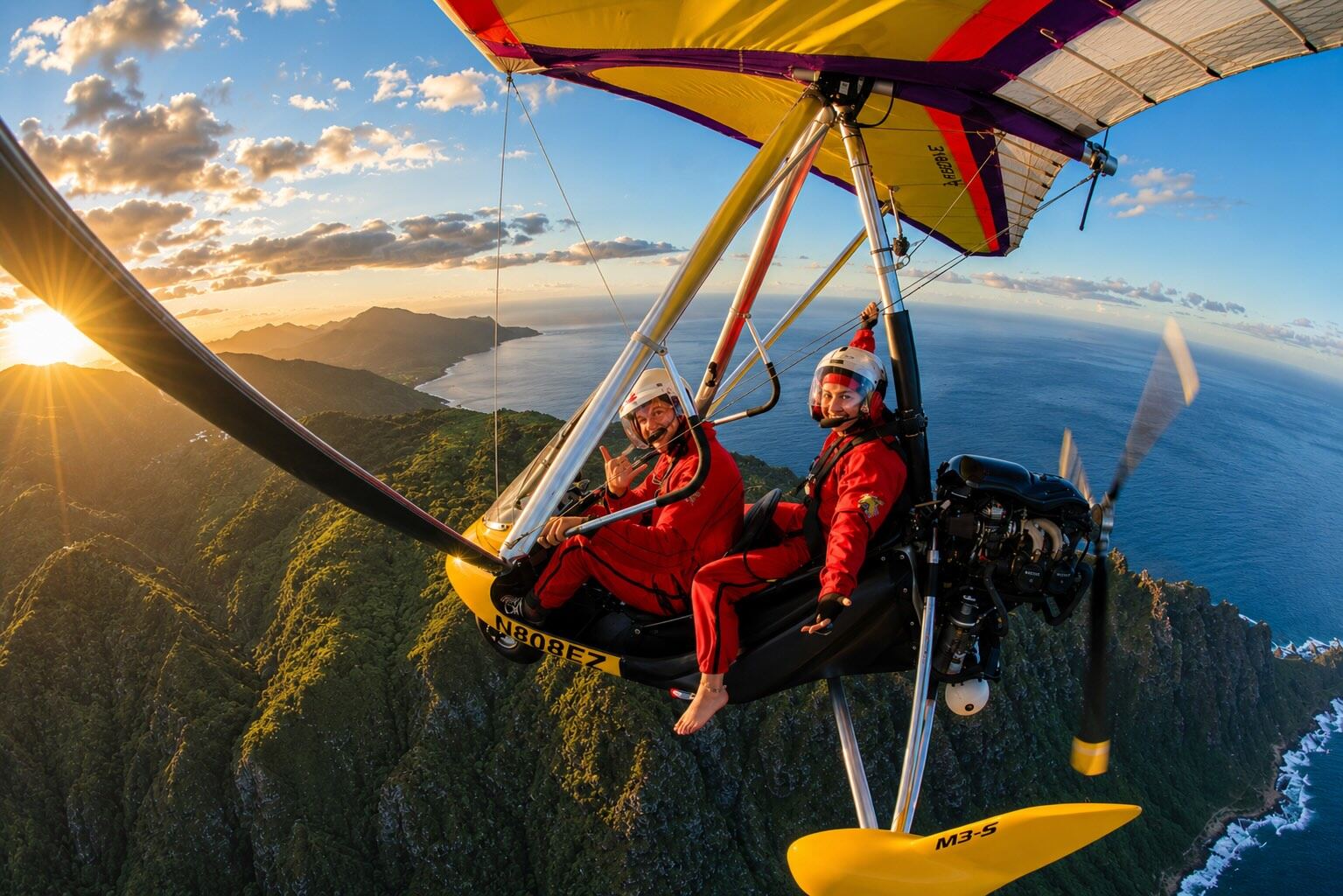 Two flyers in red flight suits aboard the trike, smiling at the camera, the sun low over green ridges and the coastline below