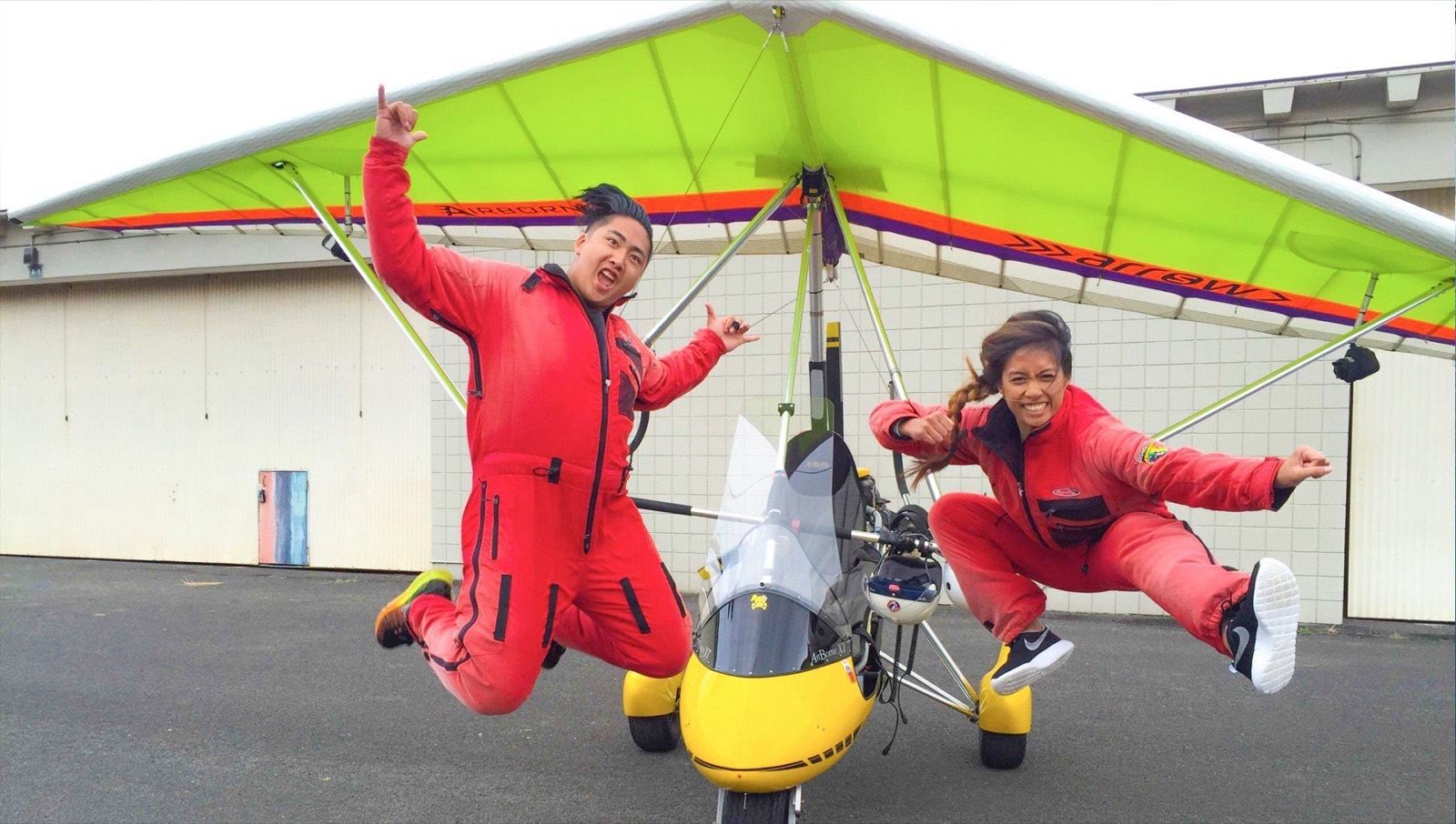 Two flyers in red flight suits leaping beside the trike’s green wing at the hangar